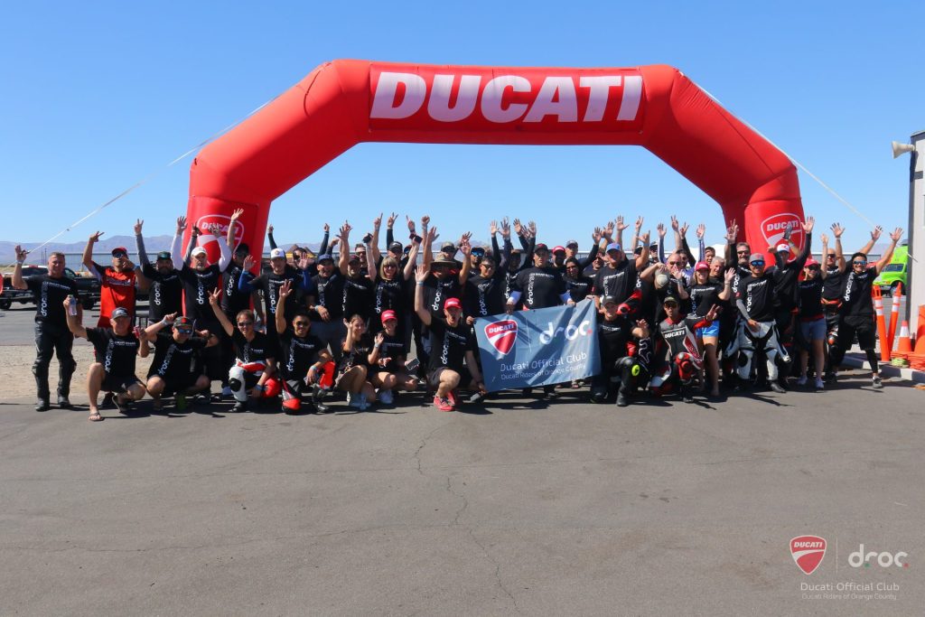 Members raise their arms in a group photo under the red Ducati inflatable arch at Chuckwalla Valley Raceway, September 2023