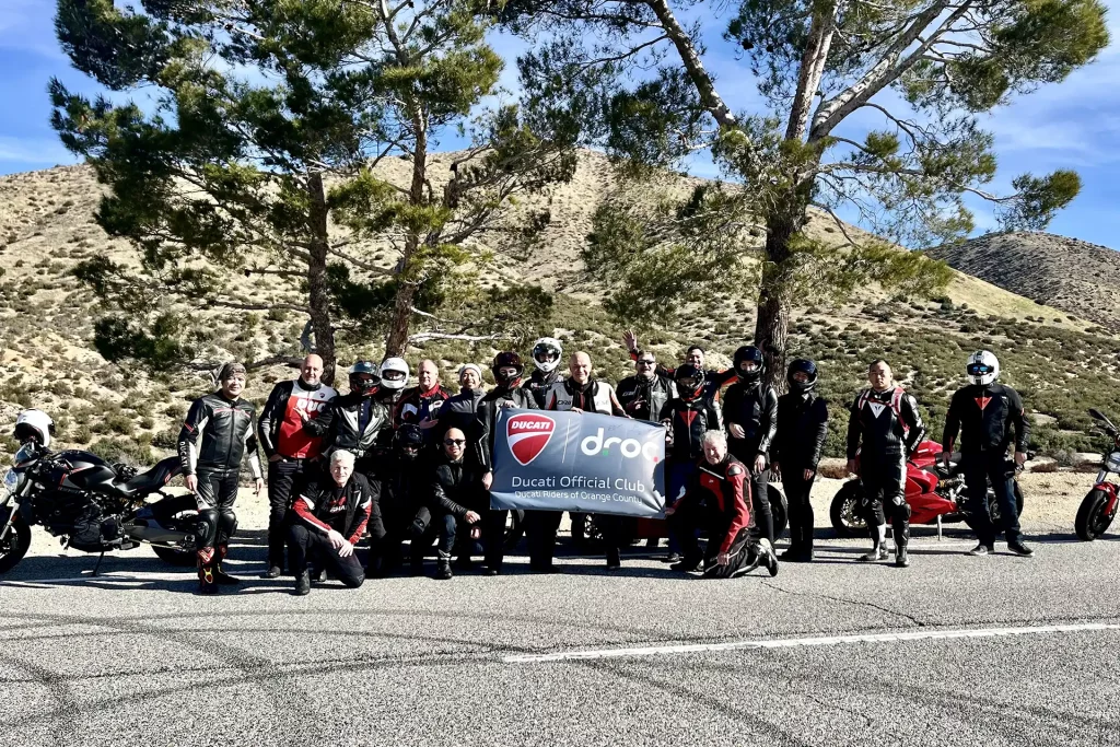 Ride with DROC: the six ways we ride 7 Riders in gear with a DROC Ducati Official Club banner on a roadside pullout below dry hills