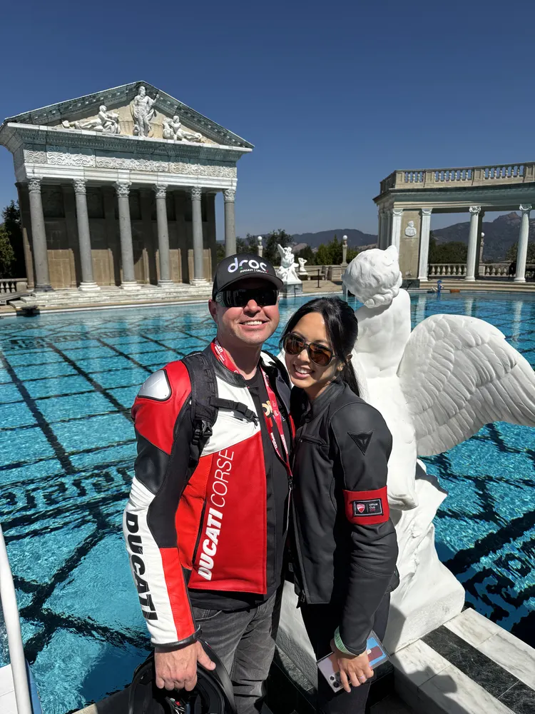 Man and woman standing together by a pool with a decorative white statue, a building with columns in the background.