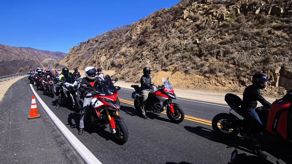 Motorcycles lined up on a road beside a rocky hillside, with riders in helmets and gear, some wearing colorful jackets.