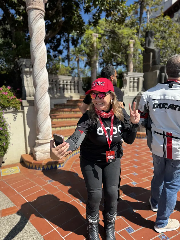 Woman wearing a black shirt with "droc" and a red cap, taking a selfie in a garden with stone columns and greenery.