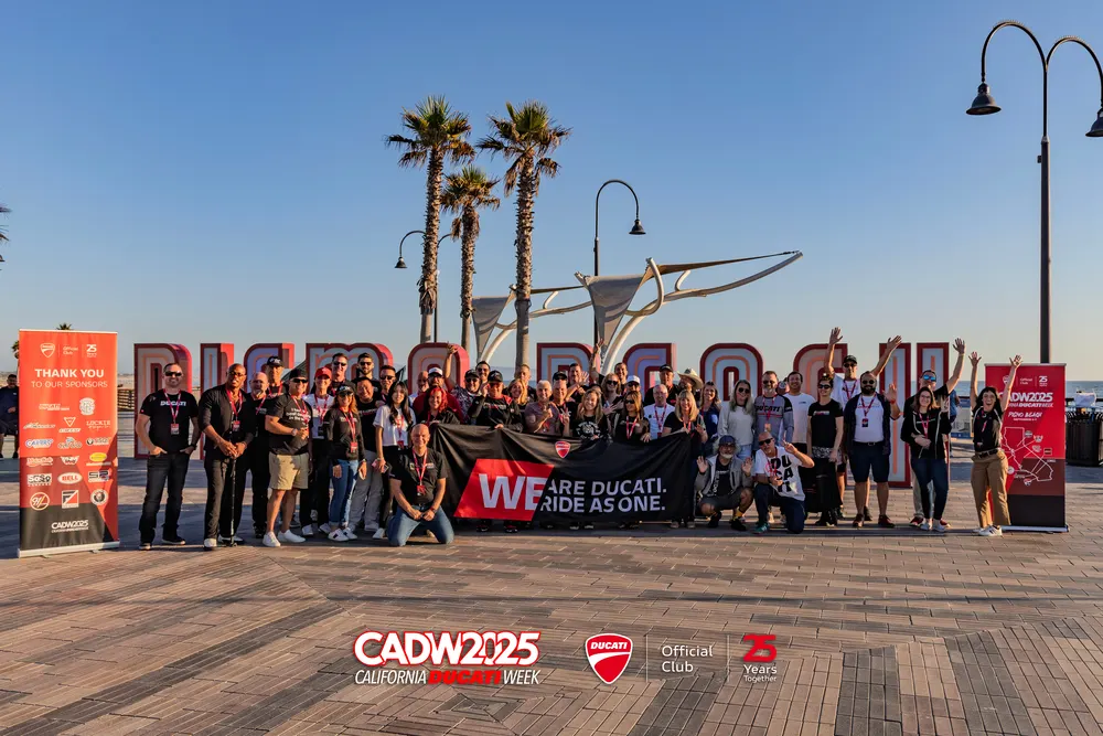 Group of people standing together, holding a banner that reads "WE ARE DUCATI. RIDE AS ONE" in front of a large sign.