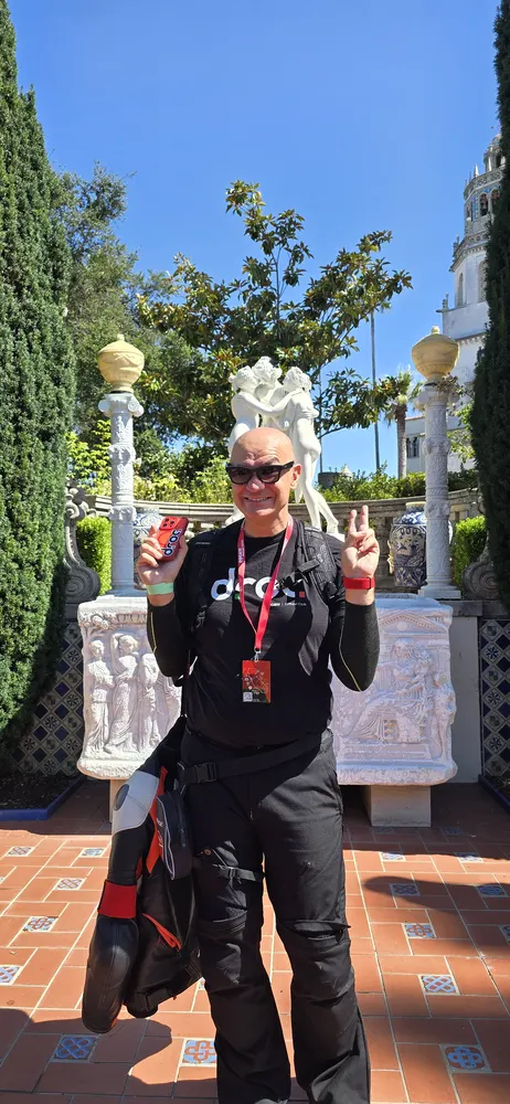 Person in black outfit with a red and black jacket holding a drink and making a peace sign in a garden with statues.