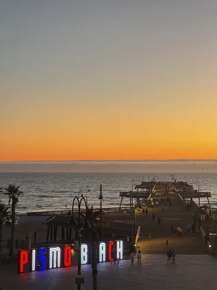 Pismo Beach sign illuminated at sunset with people walking along the pier and ocean in the background