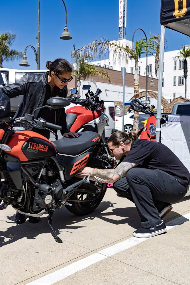 Two people working on a motorcycle labeled "SCRAMBLER" in a sunny outdoor setting with palm trees and other bikes visible.