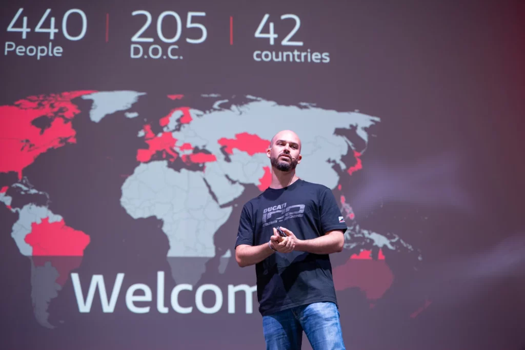 Ride with DROC: the six ways we ride 31 Speaker on stage in front of a slide reading 440 people, 205 D.O.C., 42 countries over a world map