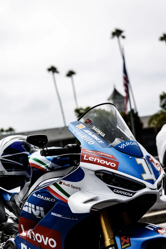 Motorcycle with blue and white bodywork displaying logos, parked with palm trees and a flag in the background
