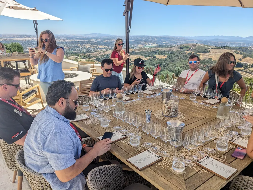Group of people seated around a large wooden table with wine glasses and menus, overlooking a landscape with hills.