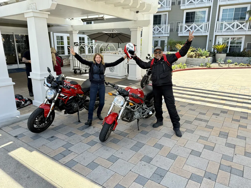 Two riders, one woman and one man, are standing beside two red motorcycles, with the woman raising her arms in excitement and the man holding a helmet, in a paved area with a building in the background.