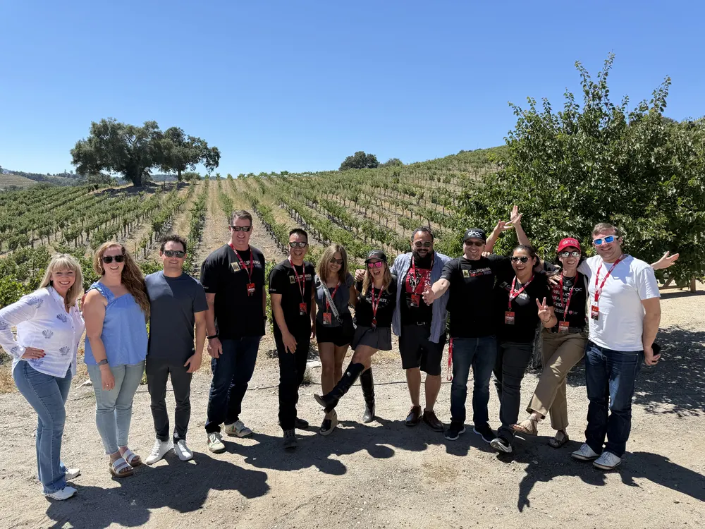 Group of eleven people posing together outdoors in a vineyard with rows of grapevines and trees in the background
