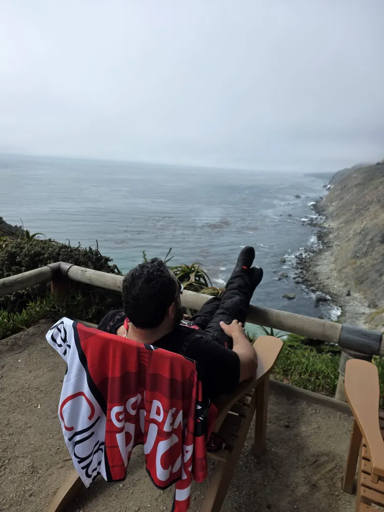 Person sitting on a chair overlooking the ocean, wearing black clothing, with a red and white towel draped over the chair.