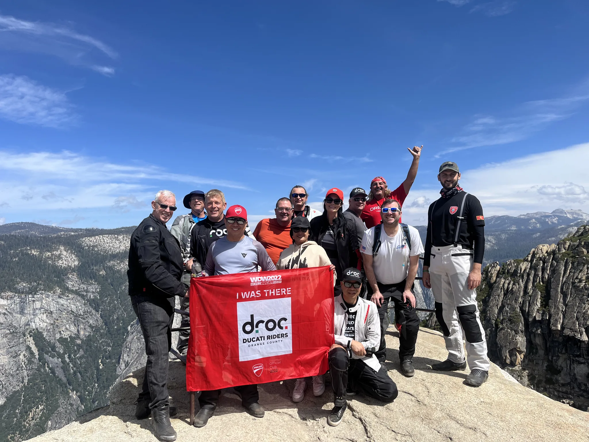 Fourteen DROC riders in gear stand on pale granite slabs at a high Yosemite overlook, holding a red DROC I Was There banner.