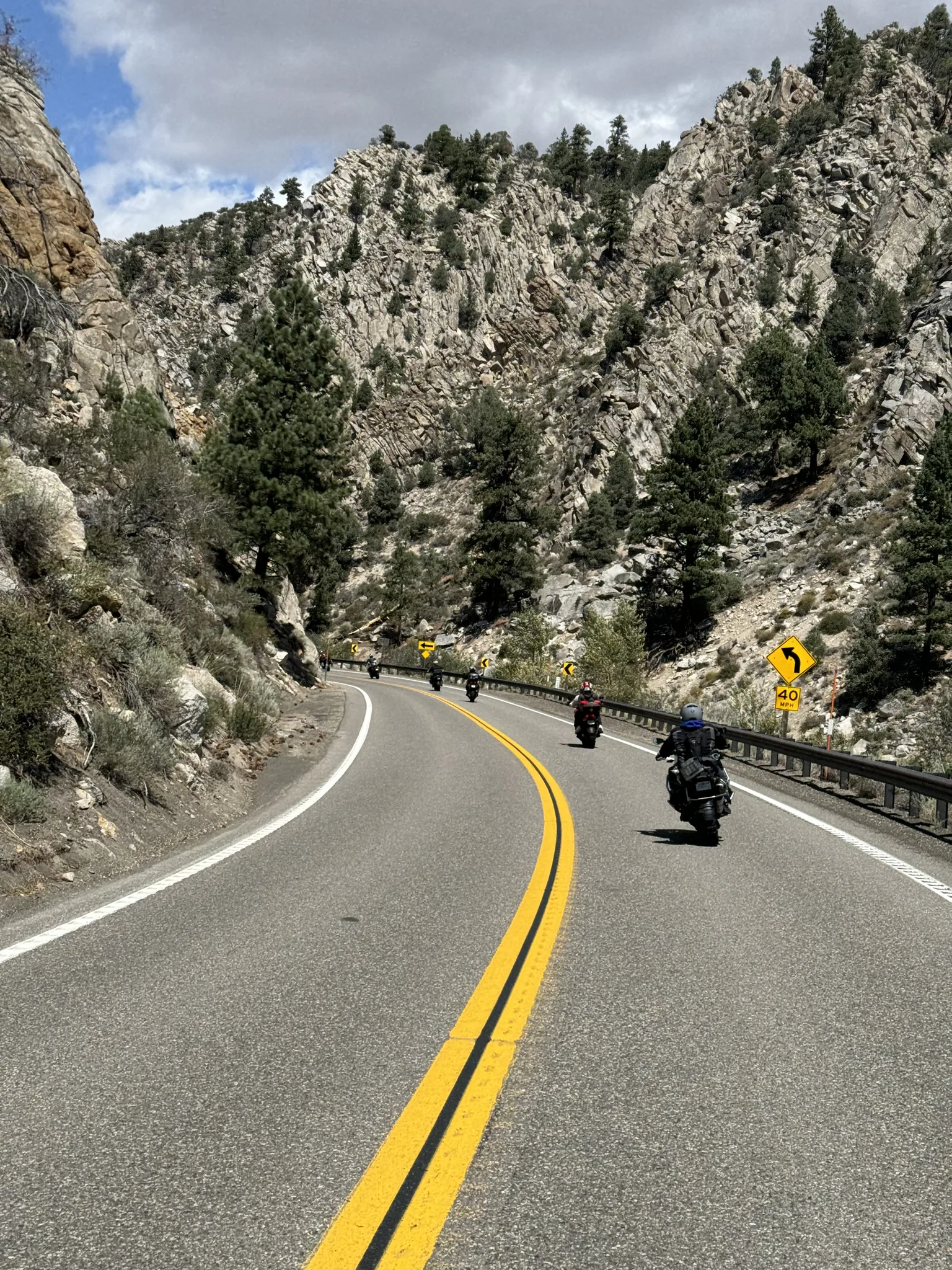 Two motorcycles come around a double-yellow curve in a granite and pine canyon on US 395, steep rock walls rising on both sides.