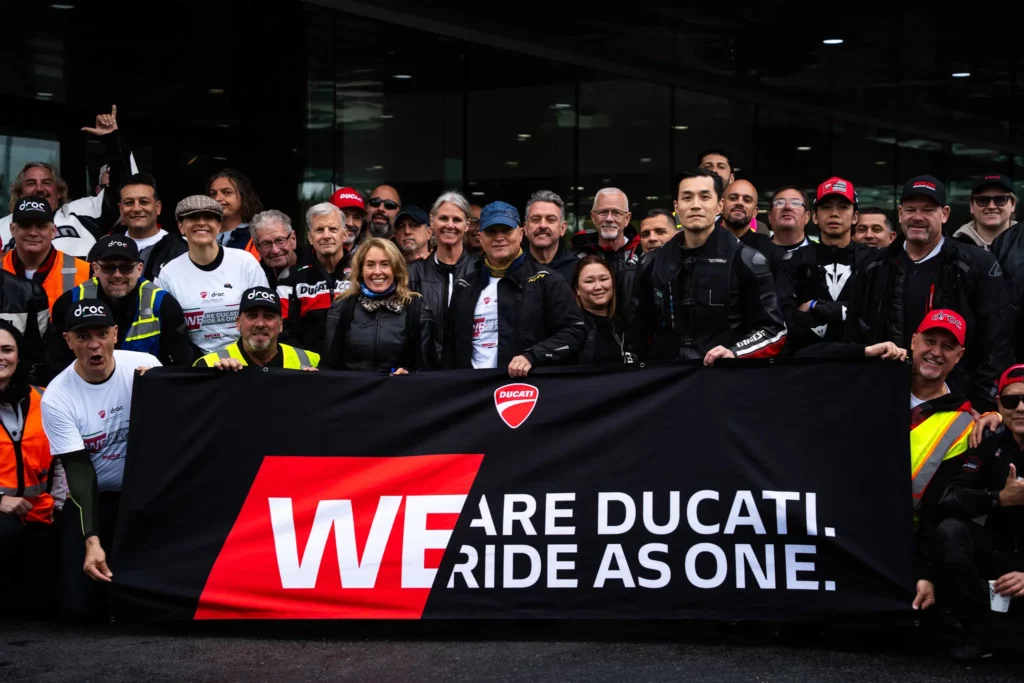 Group of people posing together, holding a banner that reads "WE ARE DUCATI. RIDE AS ONE." in front of a building.
