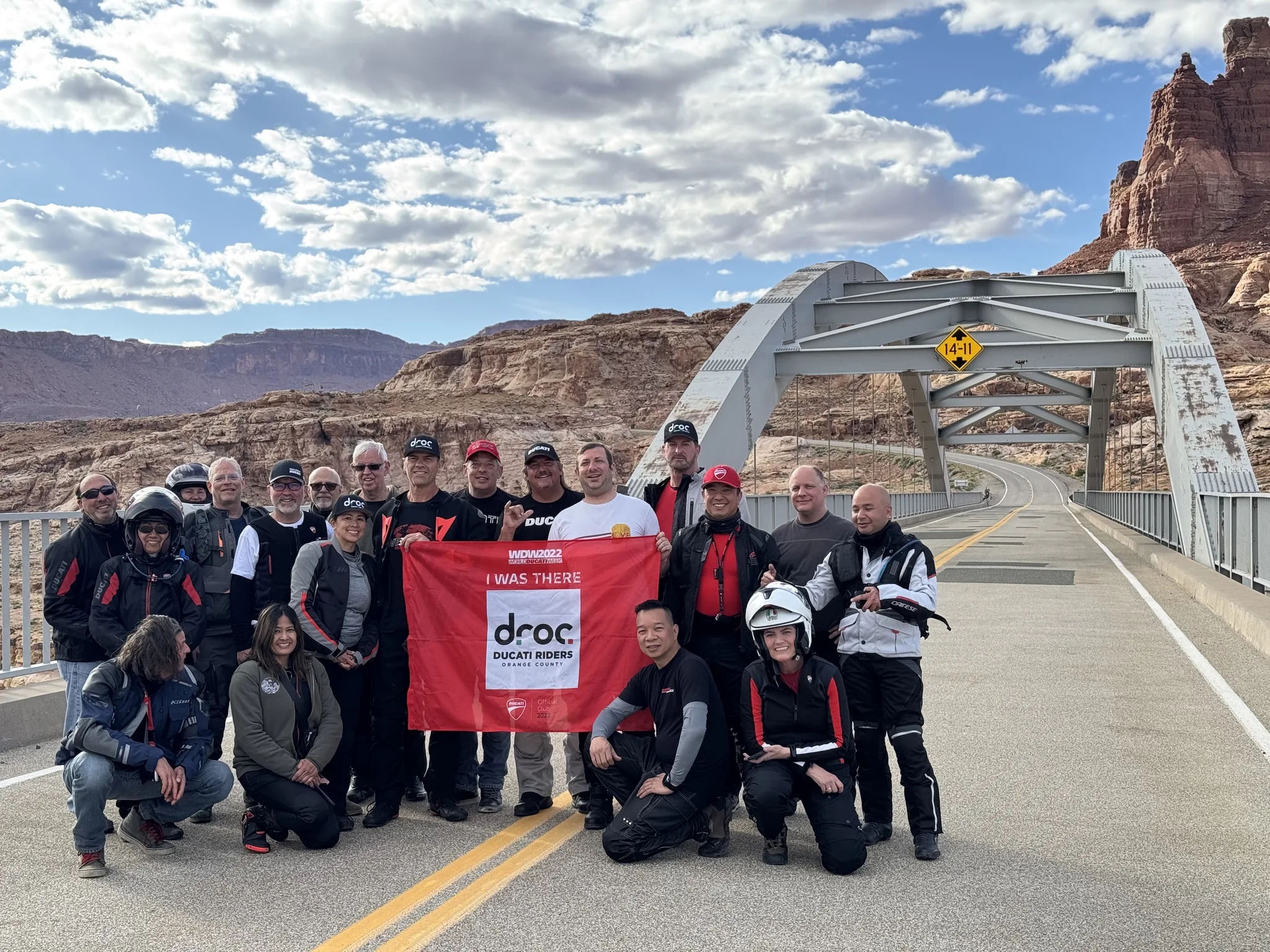 Riders stand and kneel with a red DROC banner on a steel arch bridge spanning a red rock canyon under a bright sky.