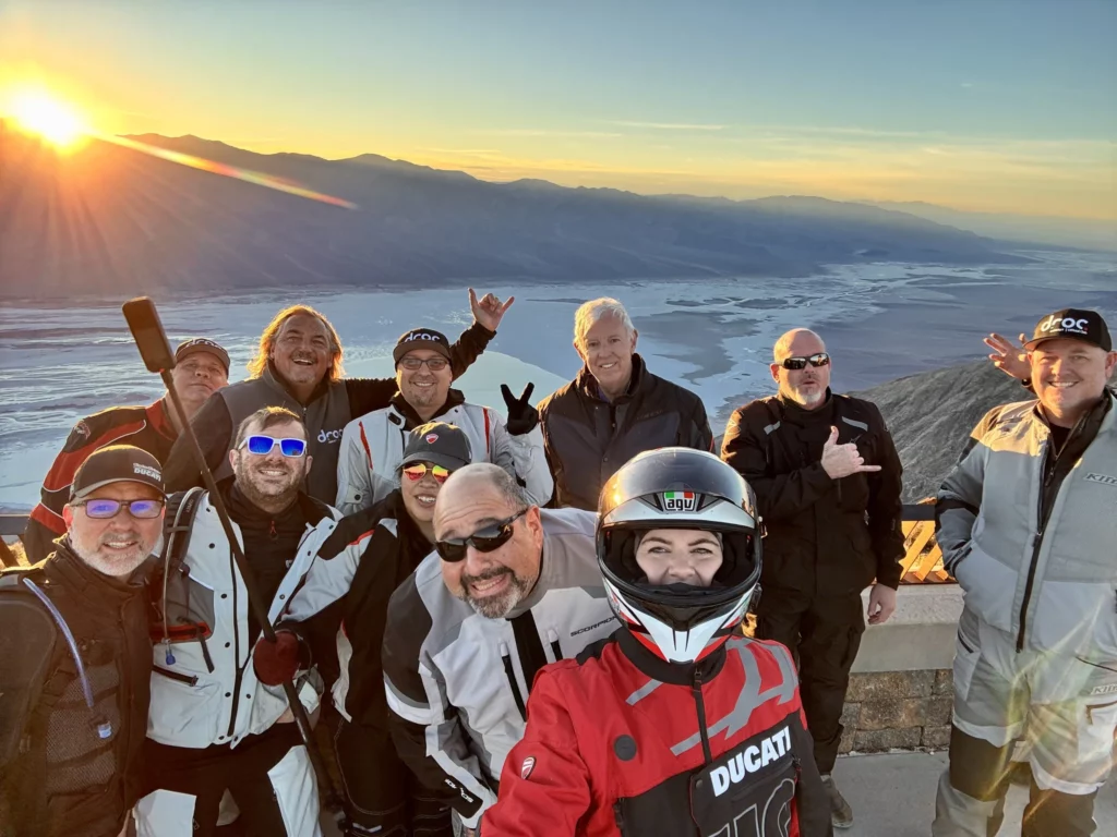 Ride with DROC: the six ways we ride 16 Nine DROC riders crowd into a sunset selfie on the rock wall at Dante's View, with Badwater Basin and the Panamint Range far below.