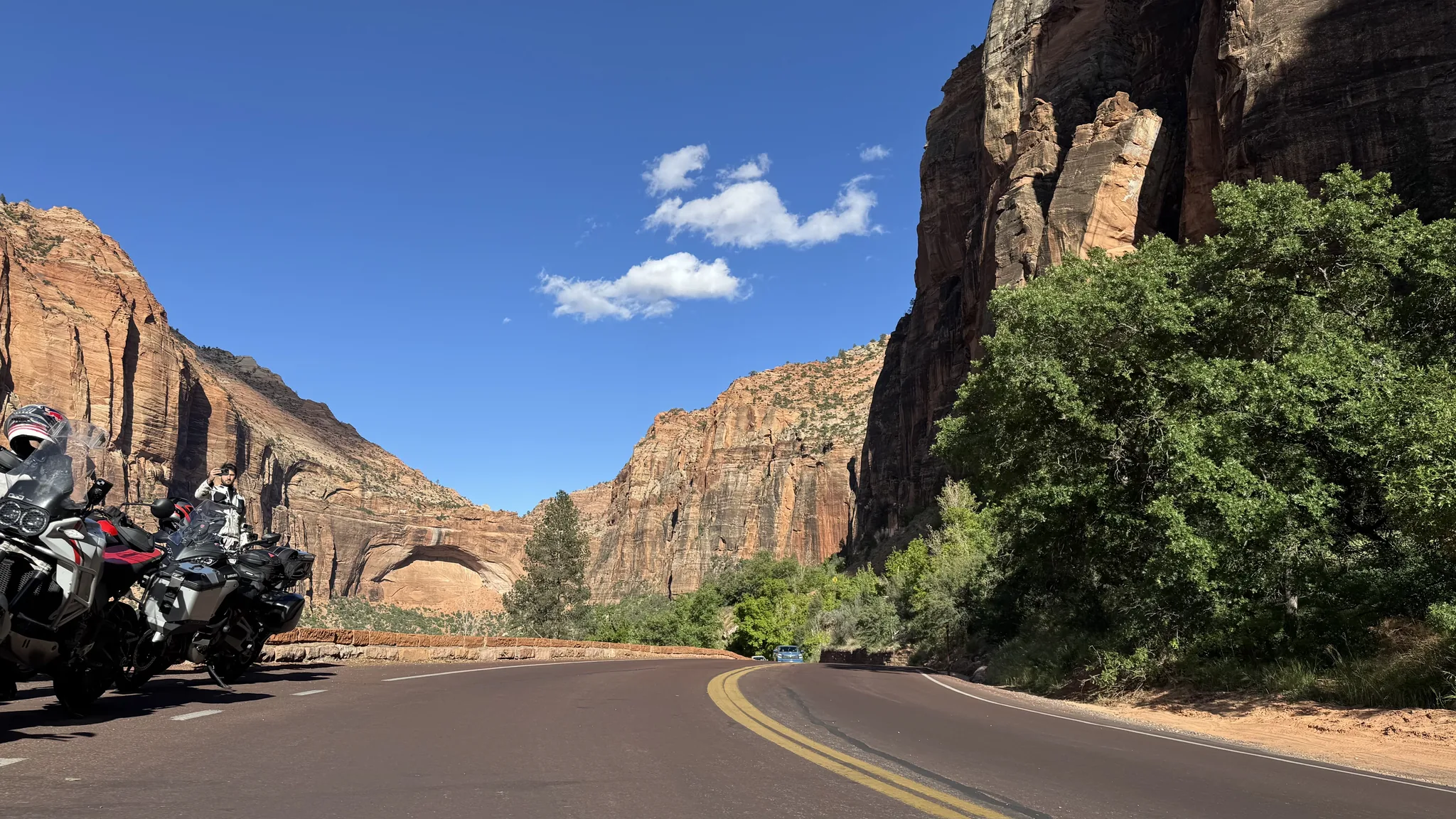 An empty two-lane road runs toward tall red and cream sandstone cliffs in Zion National Park, motorcycles parked on the left shoulder.