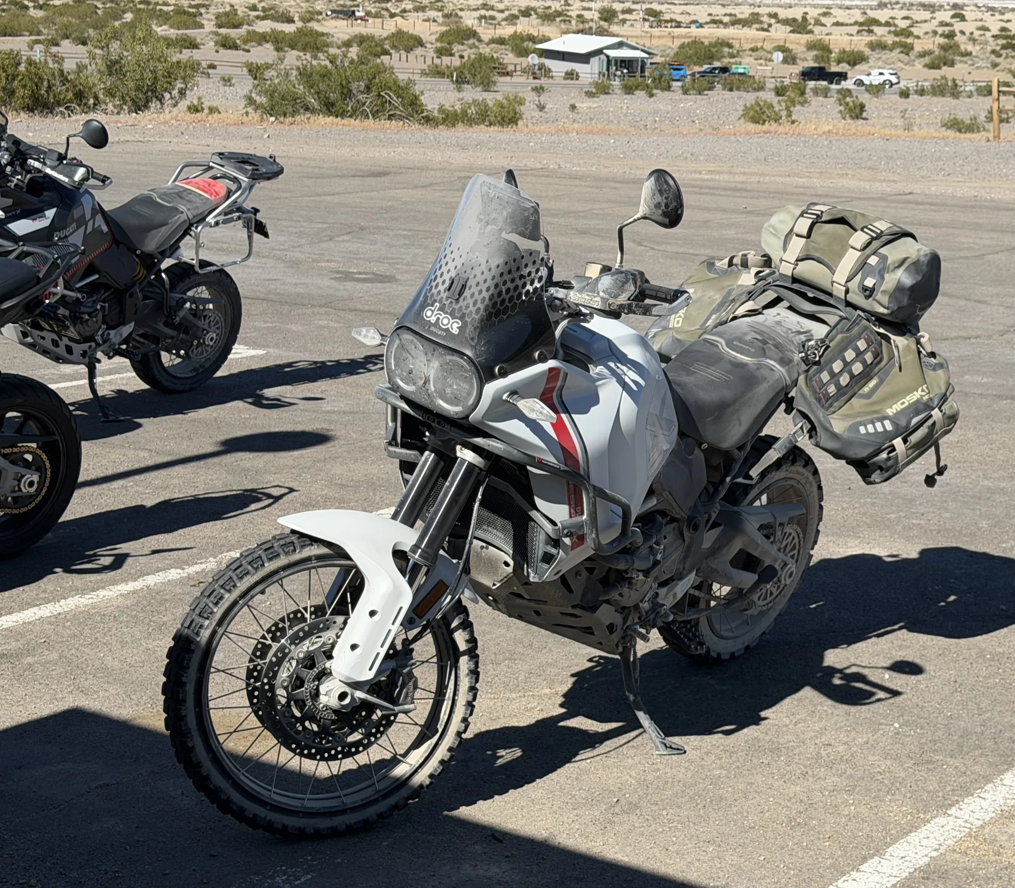 A dusty white Ducati DesertX with Mosko Moto soft luggage and knobby tires on its sidestand in a desert parking area, a Ducati Multistrada behind it.