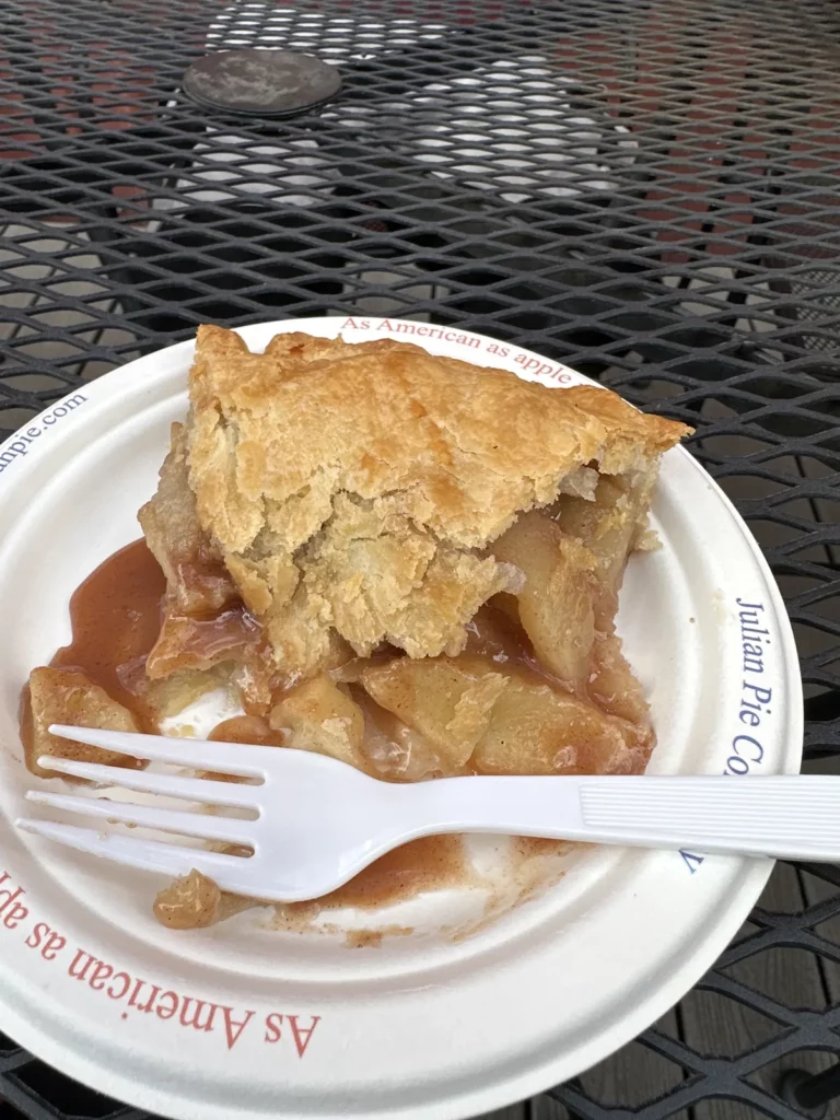 Slice of apple pie on a Julian Pie Company paper plate with a white plastic fork on a metal table