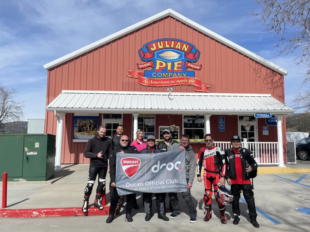 Group of nine people, some in motorcycle gear, holding a Ducati Official Club banner outside Julian Pie Company building