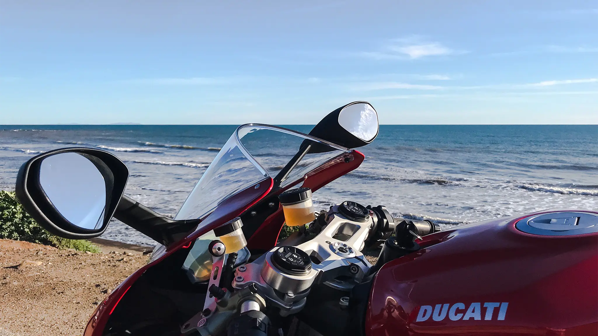 Red motorcycle parked near a beach with ocean waves in the background, visible mirrors and controls in focus