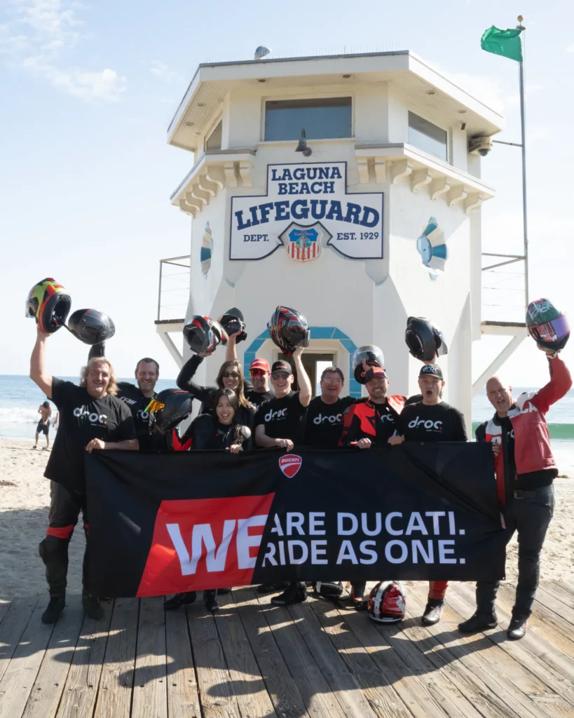 Ducati riders celebrating at Laguna Beach lifeguard tower holding a banner that says "WE RIDE AS ONE"