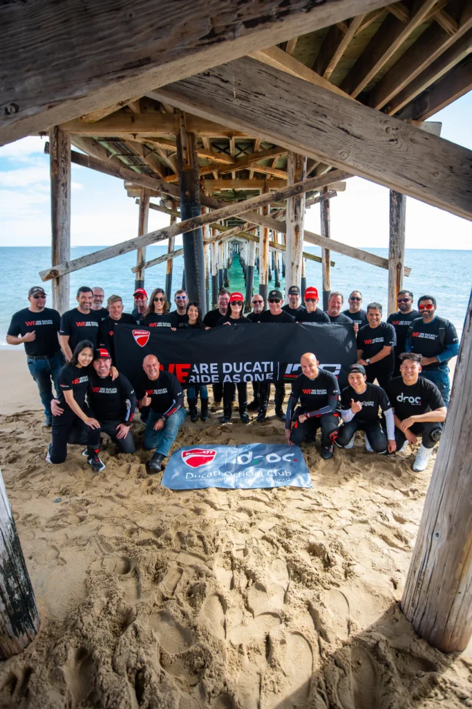 Ride with DROC: the six ways we ride 25 Group under a wooden pier holding a We Are Ducati Ride As One banner, a DROC banner on the sand