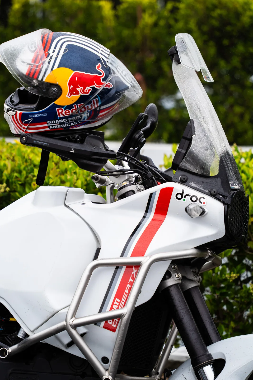 A Red Bull Grand Prix of the Americas helmet on a white DesertX wearing a DROC sticker, rain on the screen, Porsche Irvine 2025