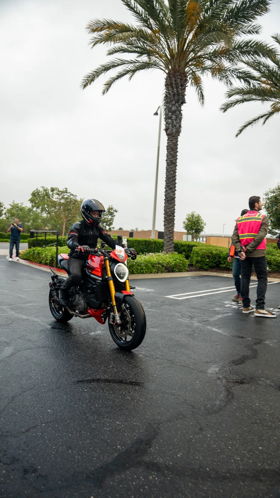 A rider on a red Monster rolls into the Porsche Irvine lot under the palms as DROC volunteers wait in pink vests, May 2025