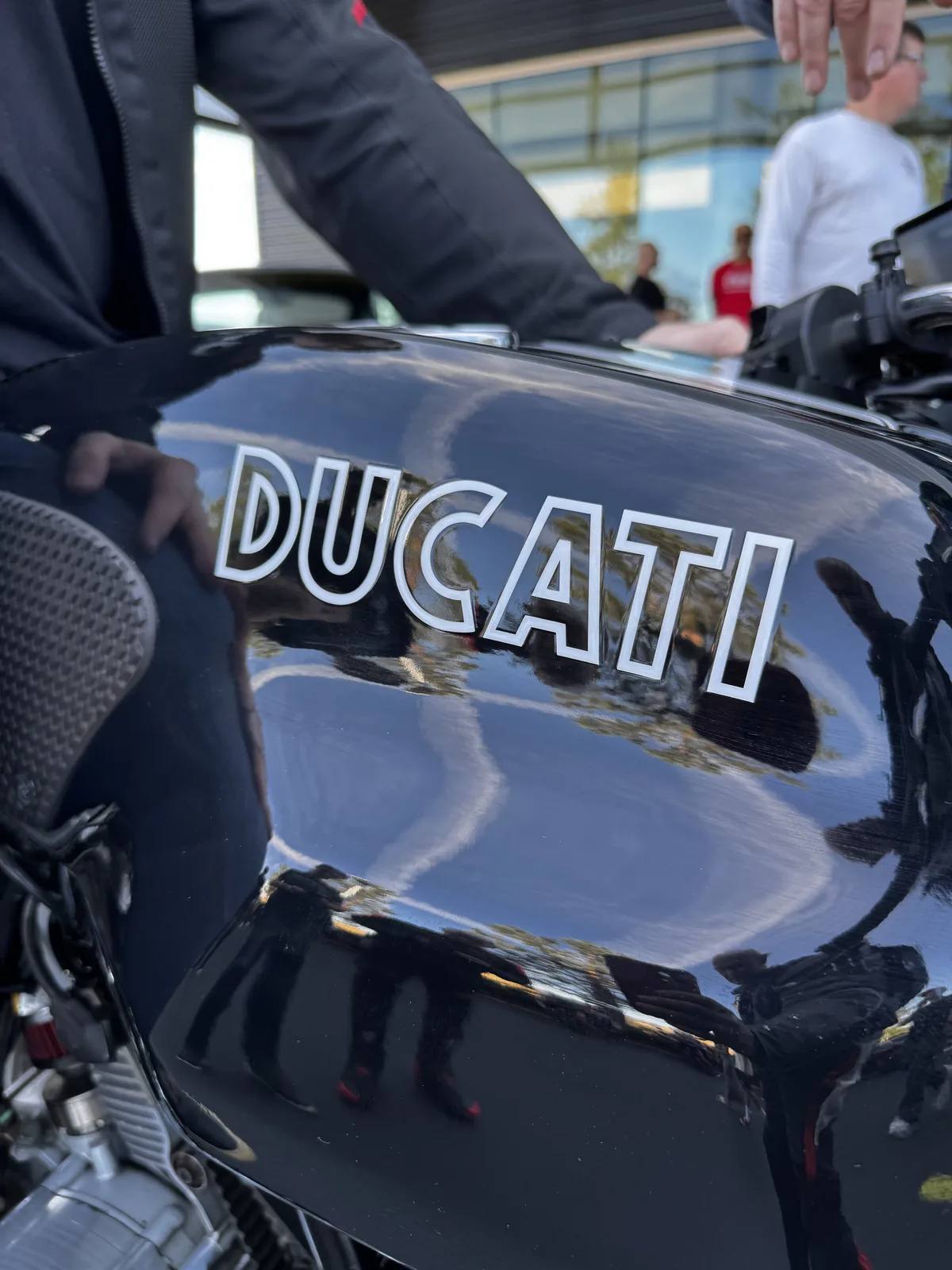 The Ducati script on a black fuel tank reflects the crowd at Porsche Irvine Cars and Coffee, March 21, 2026