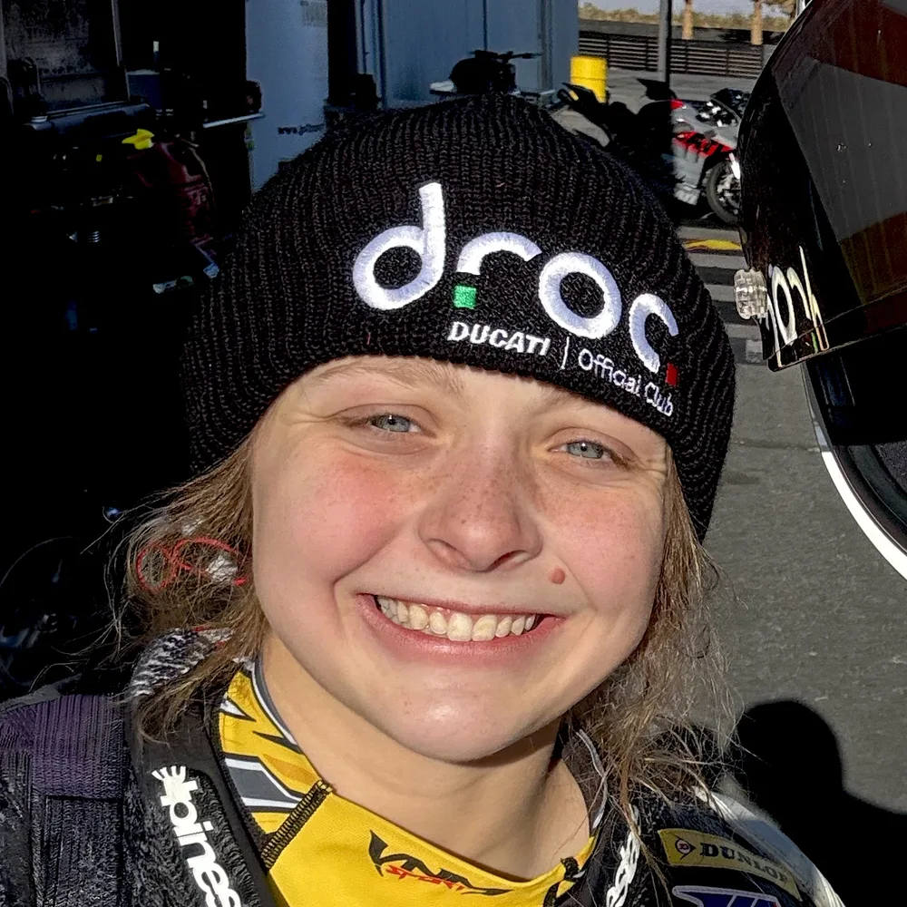 Kayla smiling in the paddock, wearing the black DROC beanie with the droc logo and Ducati Official Club embroidered in white