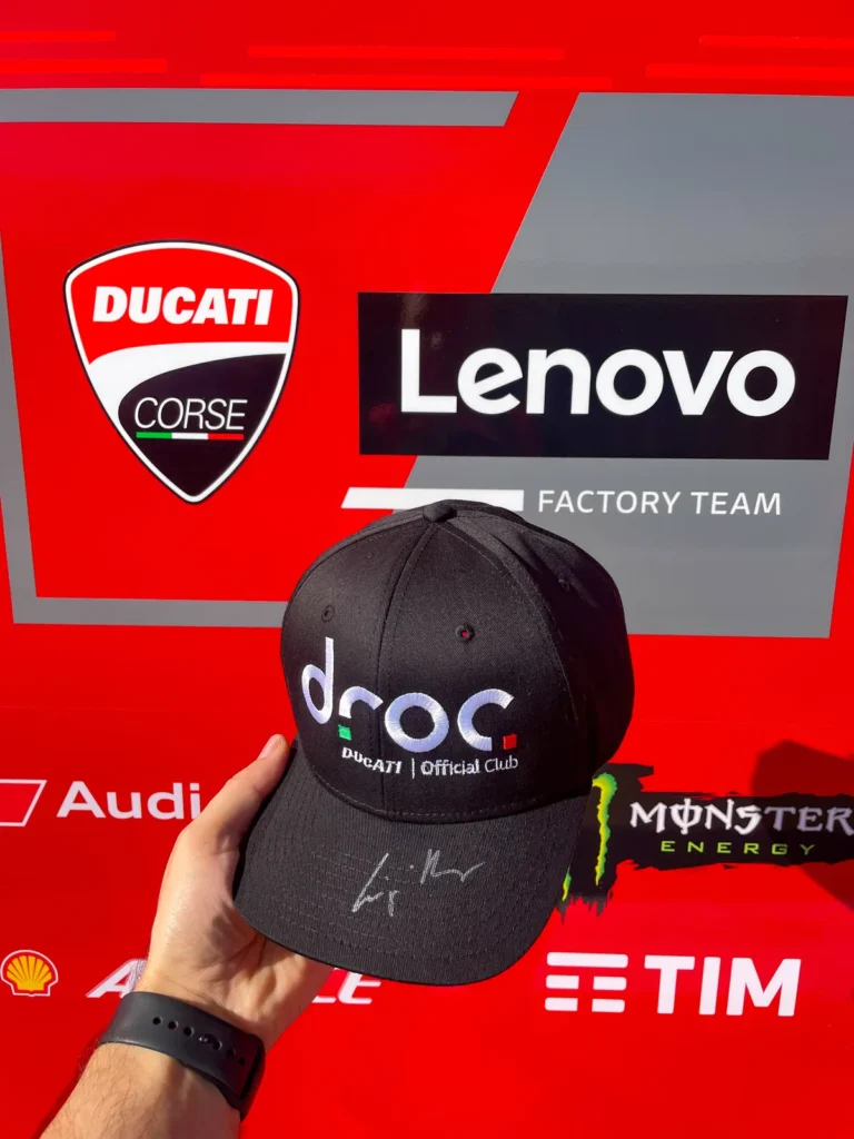 Signed black DROC cap with the Ducati Official Club logo, held up in front of the Ducati Corse and Lenovo team wall at COTA