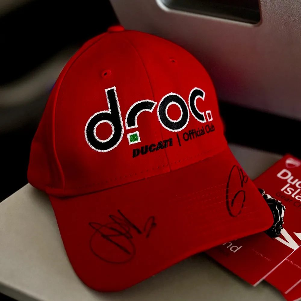 Signed red DROC cap with the Ducati Official Club logo on a tray table beside Ducati Island gear check tickets