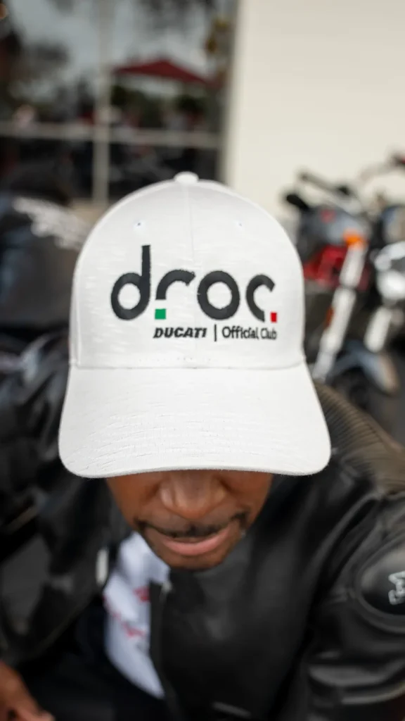 Member wearing a white DROC cap with the Ducati Official Club logo, motorcycles parked behind