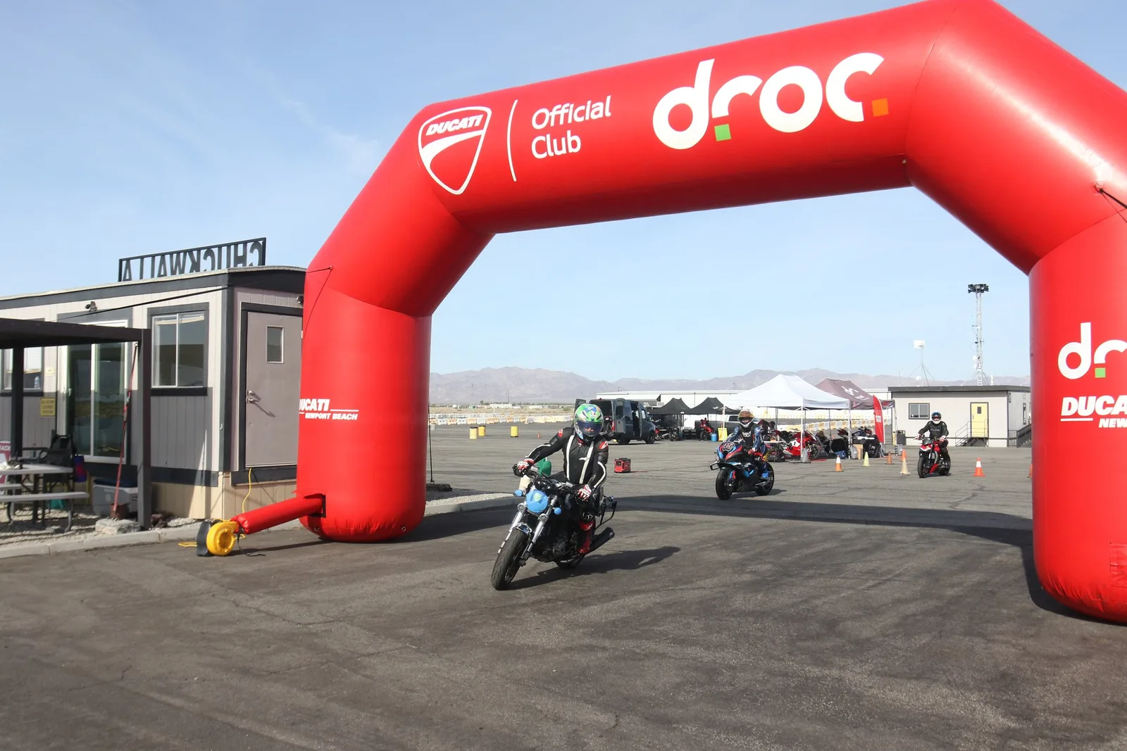 A rider in a green helmet rolls out under the red DROC Official Club inflatable arch at Chuckwalla Valley Raceway, with desert mountains behind, March 2026