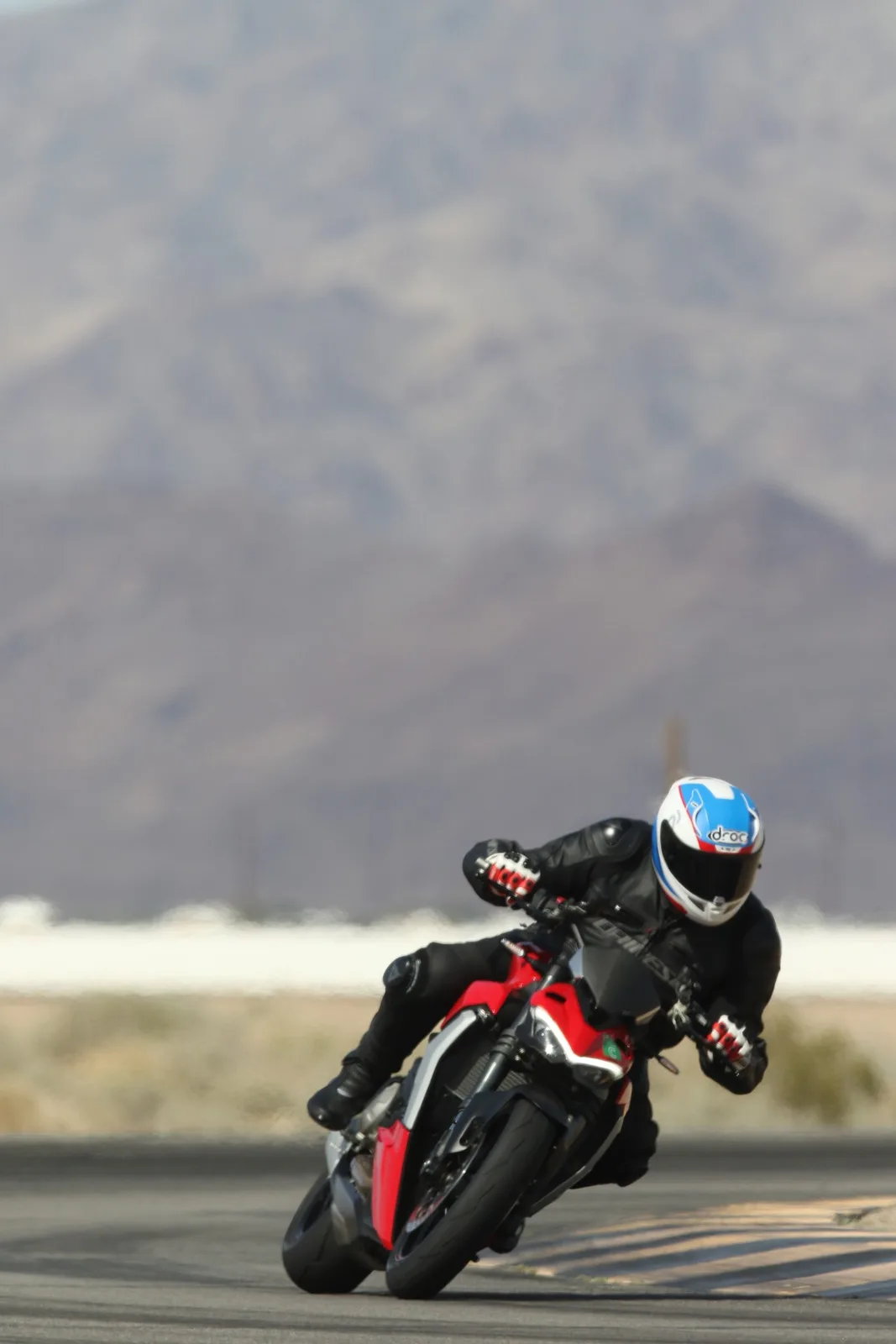 A rider in a white, blue and red helmet with a DROC logo leans a red and black Ducati through a corner at Chuckwalla Valley Raceway, March 2026