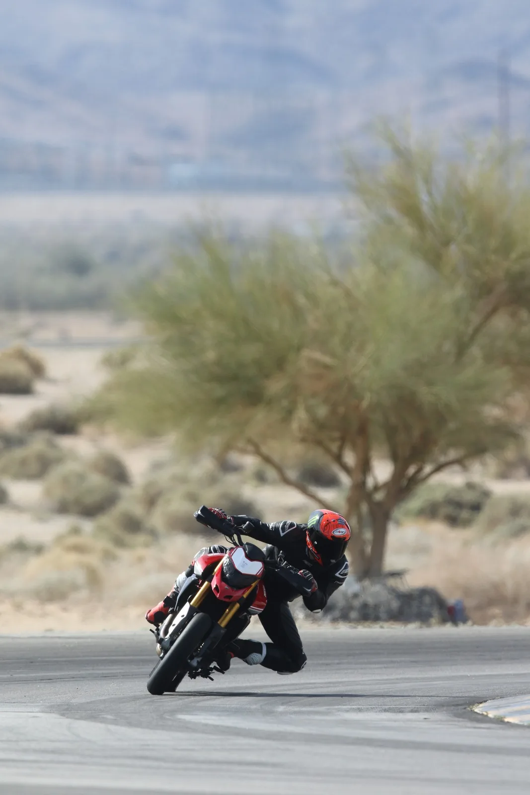 A rider in a red helmet leans a red Ducati hard into a corner on the Chuckwalla Valley Raceway track, with a desert tree and hazy hills behind, March 2026