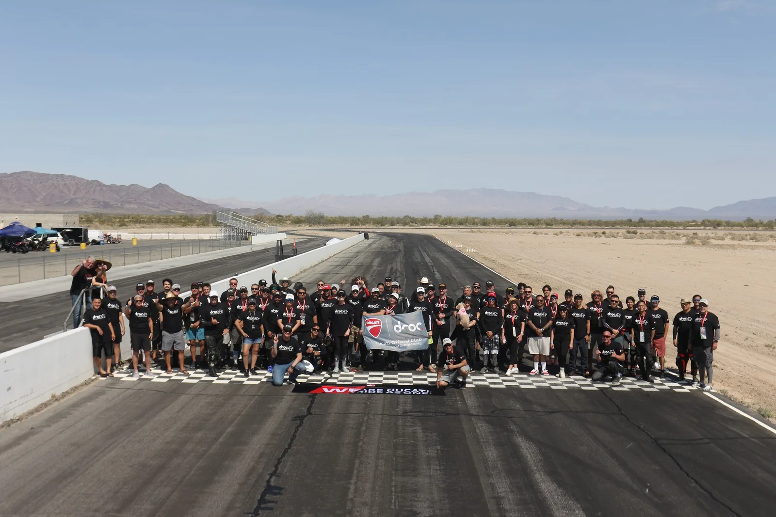 The DROC track day group in black shirts gathers behind a club banner on the finish line at Chuckwalla Valley Raceway, with desert mountains behind, March 2026