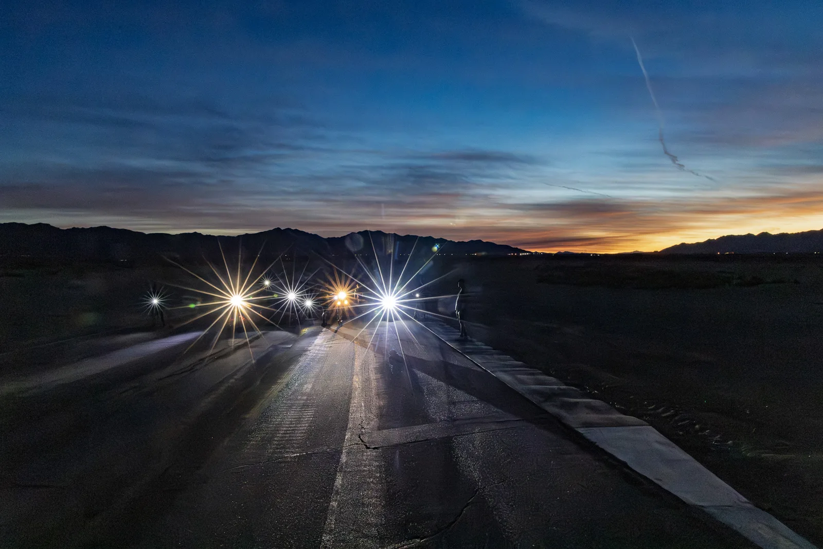 Motorcycle headlights glow in starbursts on the track at Chuckwalla Valley Raceway under an orange and blue desert sunset, November 2025