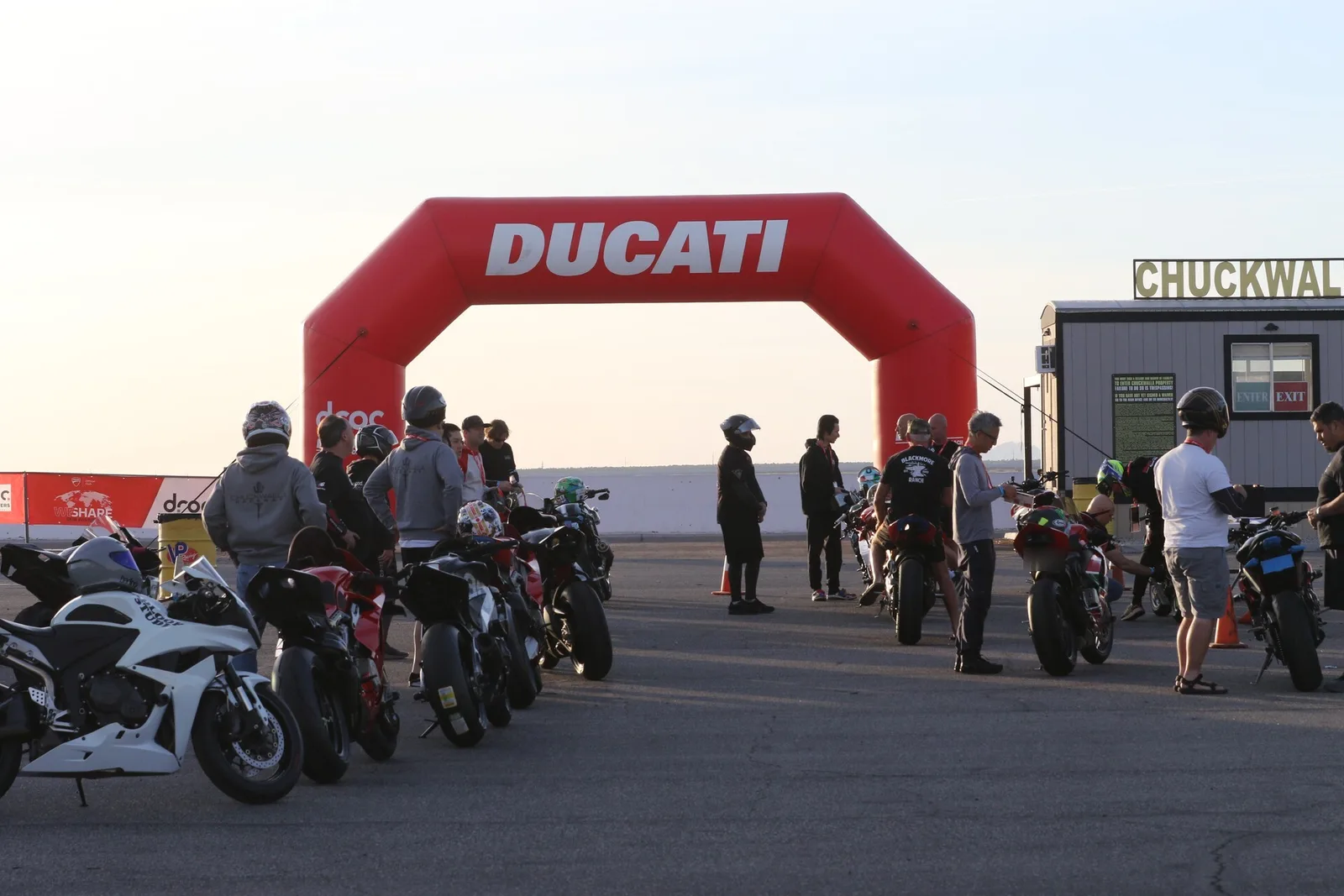 Riders in helmets and hoodies stand among parked motorcycles beside the red Ducati inflatable arch at first light at Chuckwalla Valley Raceway, March 2026