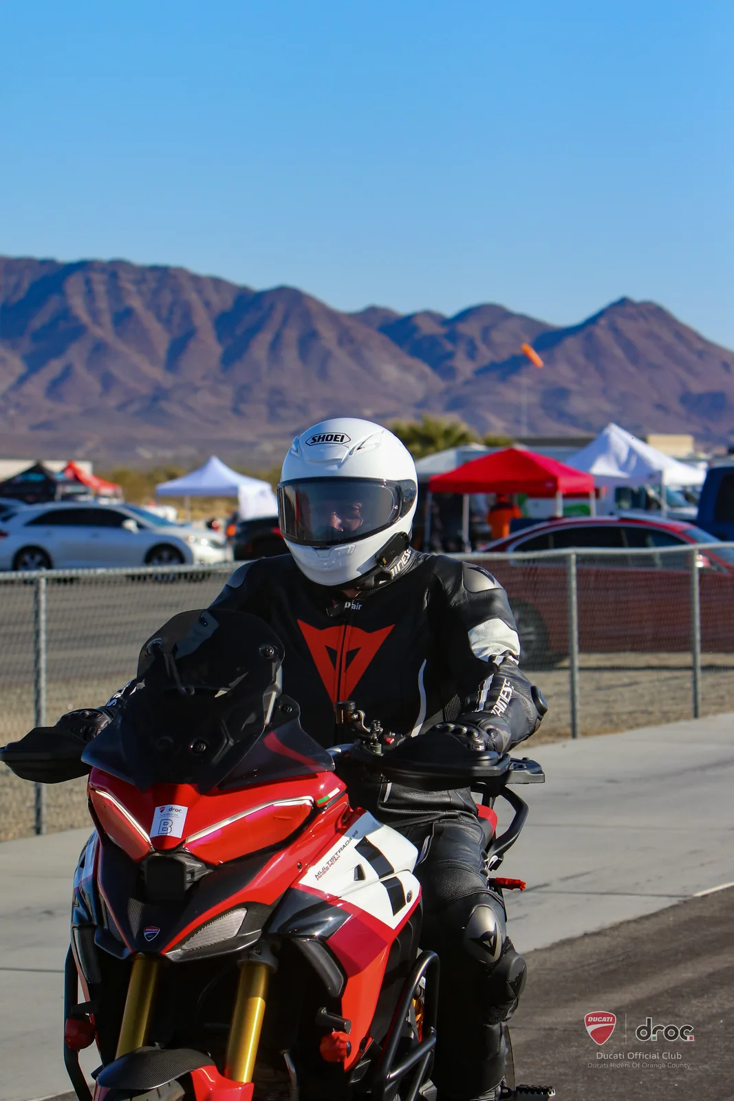 A rider in a white helmet on a Ducati Multistrada V4 Pikes Peak in the pit lane at Chuckwalla Valley Raceway with desert mountains and paddock tents behind, March 2025