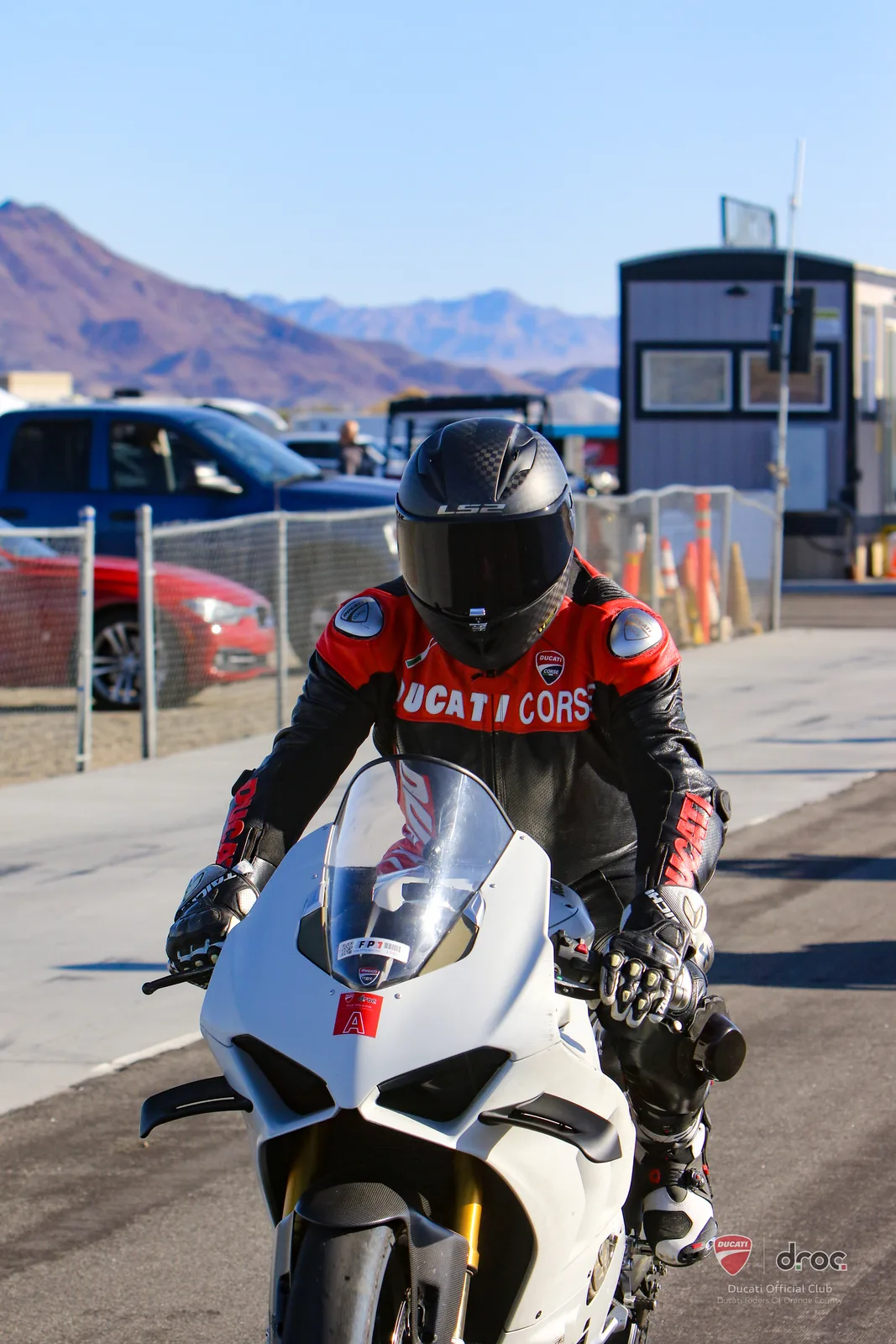A rider in Ducati Corse leathers and a black helmet rolls down the pit lane on a white Panigale at Chuckwalla Valley Raceway, March 2025