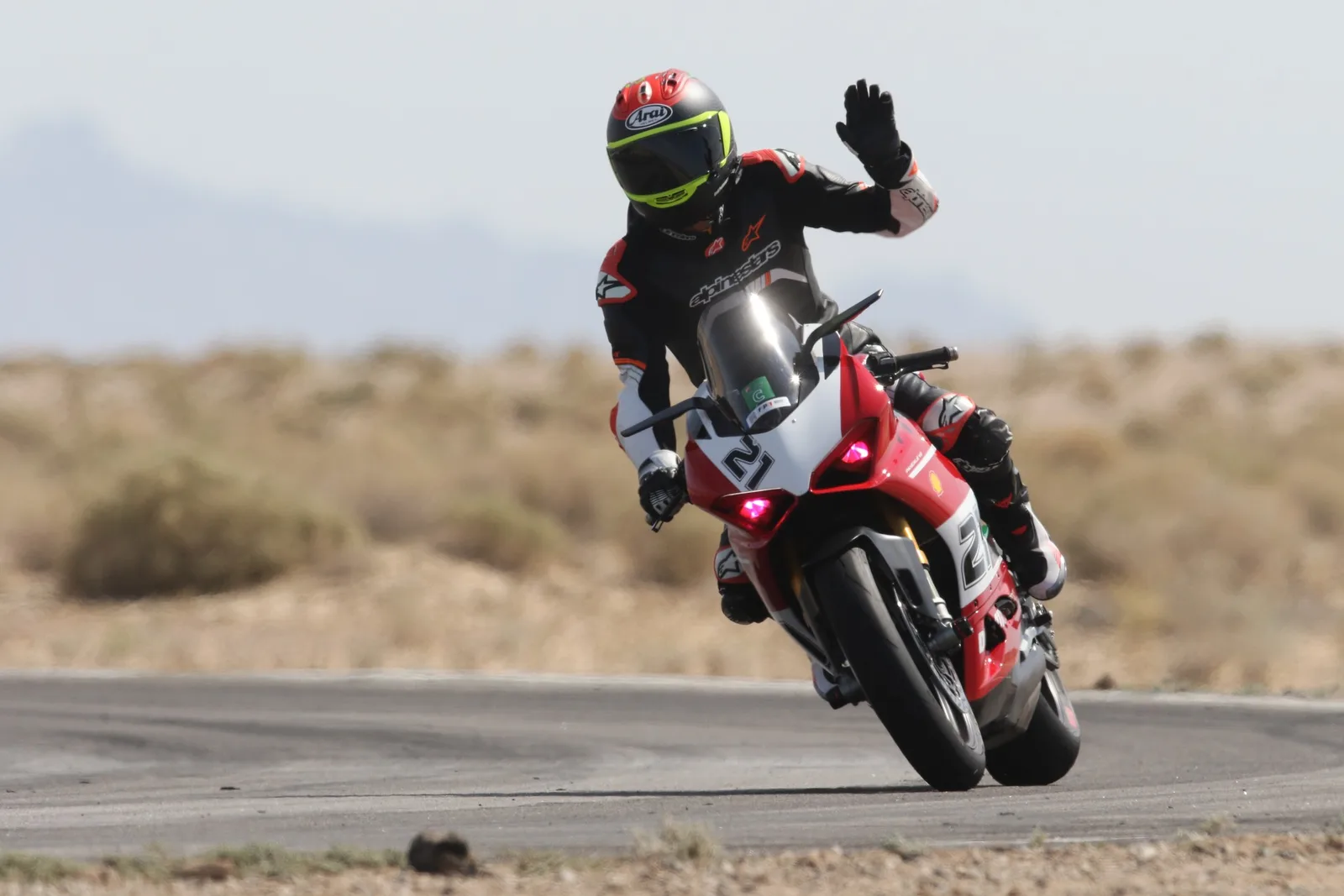 A rider on a red and white Ducati Panigale number 21 raises a gloved hand to wave while leaning through a corner at Chuckwalla Valley Raceway, March 2026