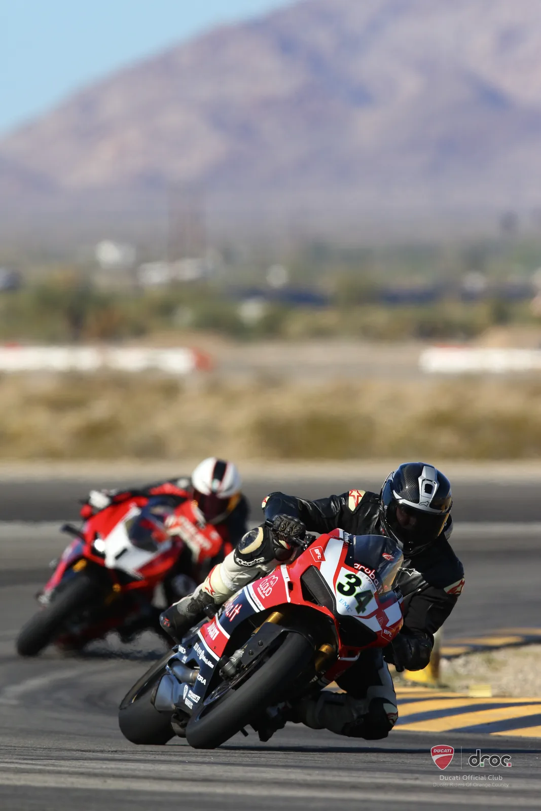 A Ducati Panigale number 34 leans through Turn 11 at Chuckwalla Valley Raceway with another rider behind and a desert mountain in the distance, March 2025