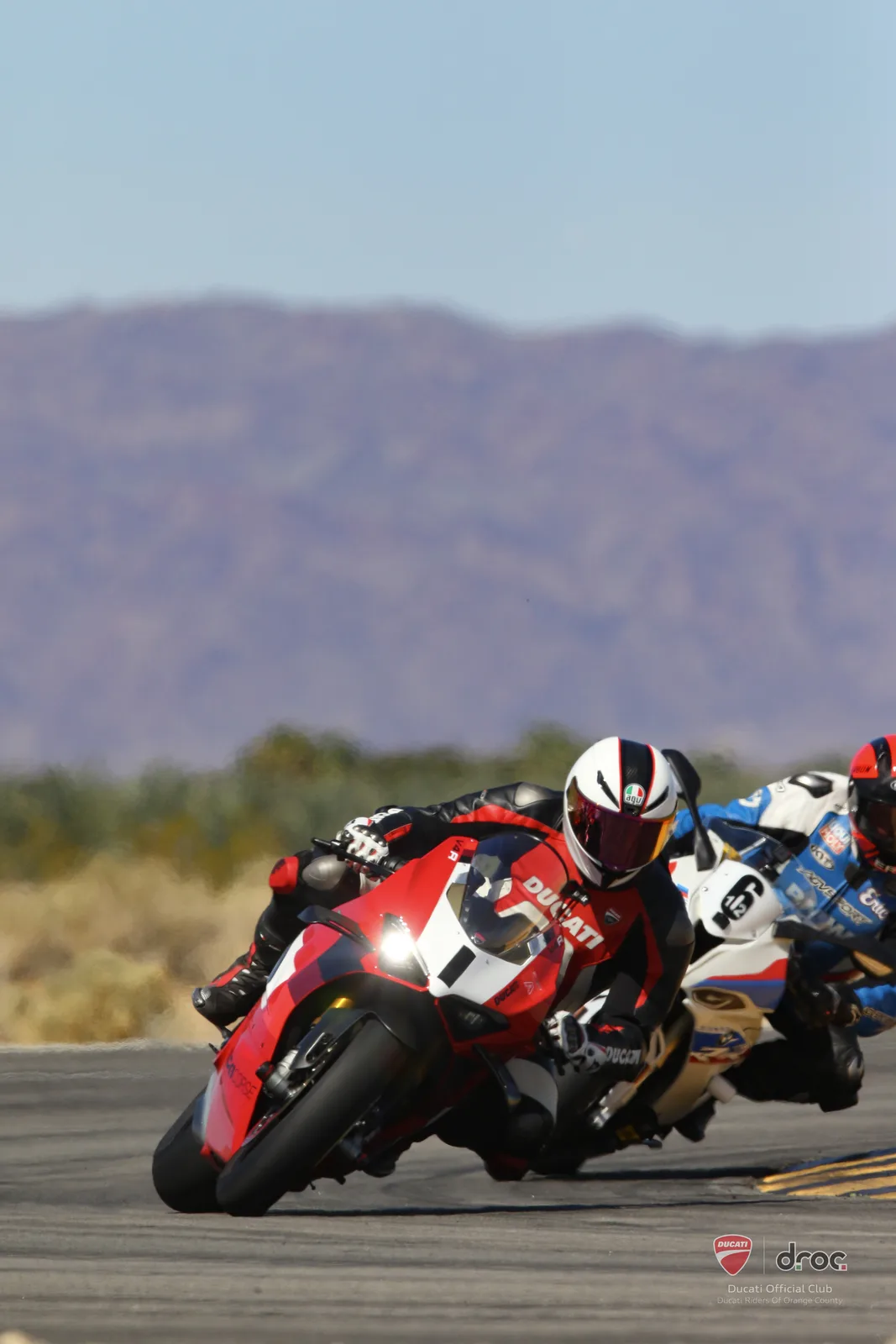 A rider in a white helmet on a red Ducati Panigale V4 R leans through Turn 16 at Chuckwalla Valley Raceway with a rider on a white and blue bike close behind, March 2025