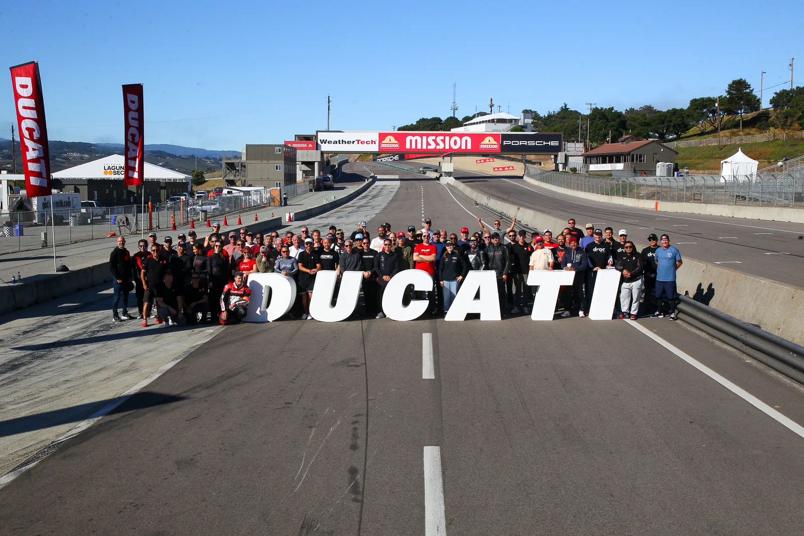 A large group of Ducati riders and staff pose behind giant white DUCATI letters on the pit straight at Laguna Seca, July 2025
