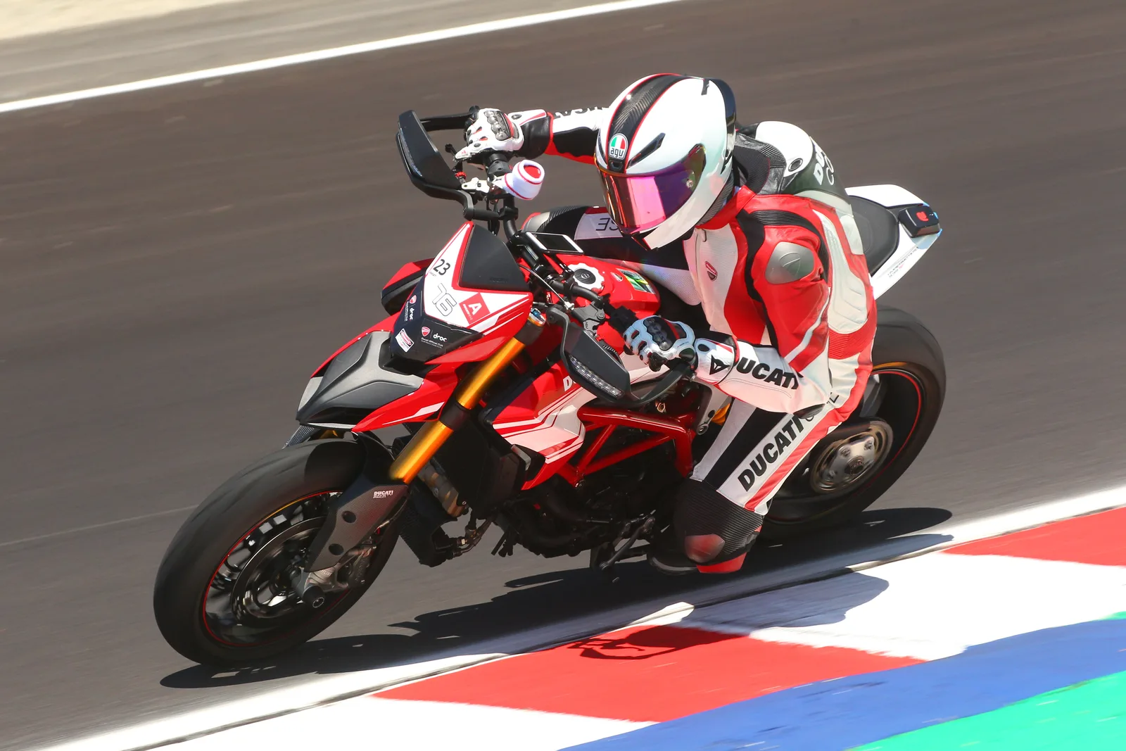 A rider in red and white leathers leans a Ducati Hypermotard with a DROC Ducati Official Club number plate through a turn at Laguna Seca, July 2023