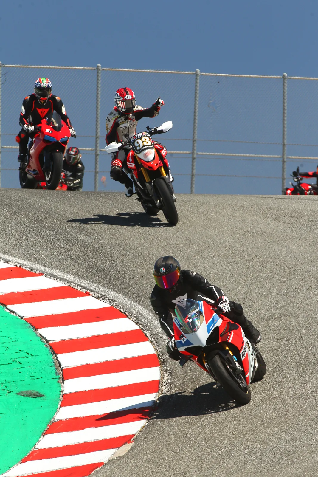 A Ducati Panigale leans through red and white curbing at Laguna Seca while a rider on a number 93 Hypermotard points ahead as more riders crest the rise behind, July 2022