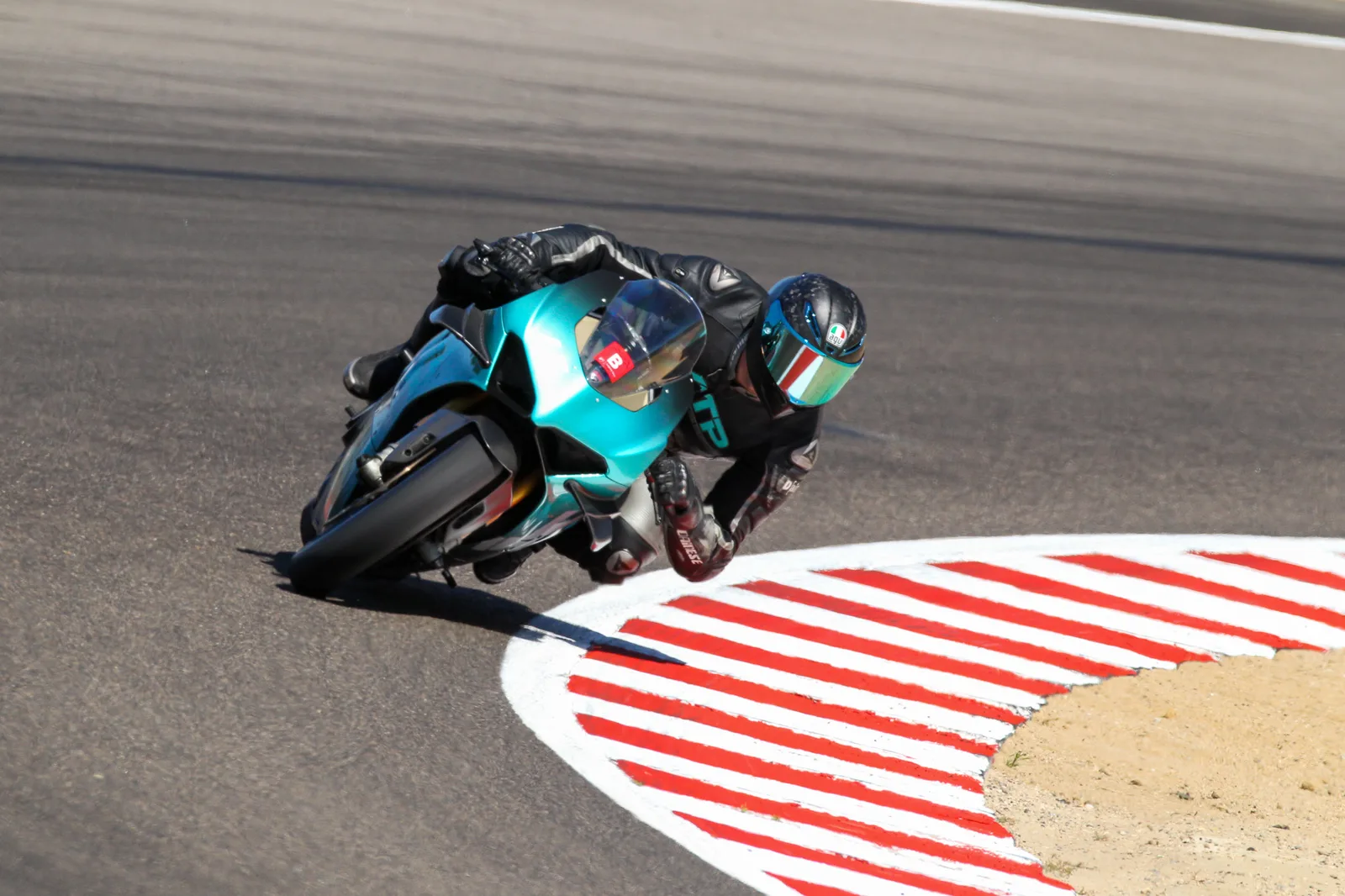 A rider in black leathers and a mirrored helmet leans a teal Ducati Panigale over the red and white curbing at Laguna Seca, July 2025