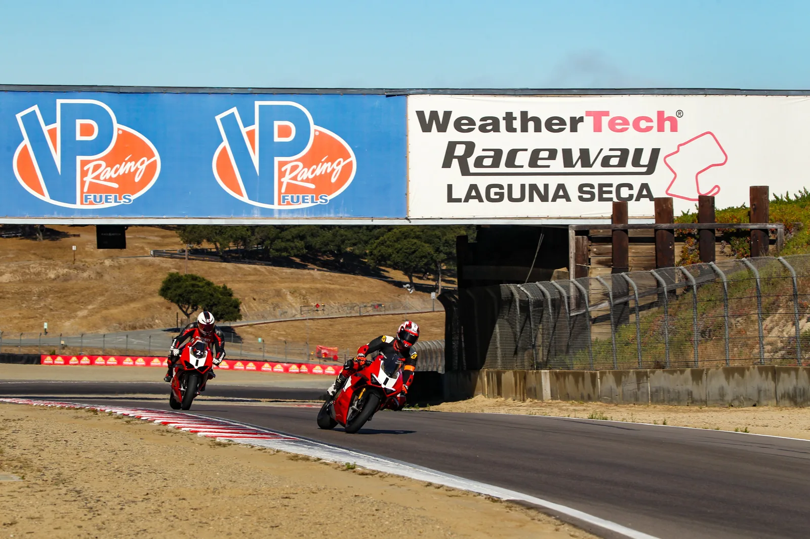 Two Ducati Panigale V4 riders lean through a corner beneath the WeatherTech Raceway Laguna Seca banner at the Ducati track day, July 2025
